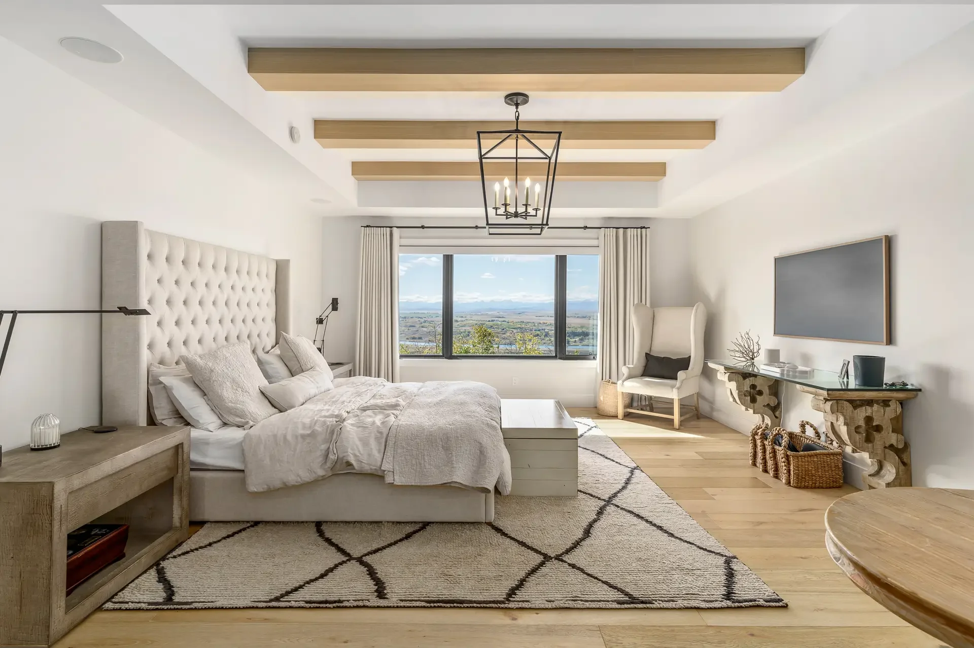 Modern bedroom with white walls, large window with scenic view, beige bed with white and gray bedding, tufted beige headboard, two bedside lamps, white armchair, decorative console table, chandelier, geometric rug, and wooden flooring.