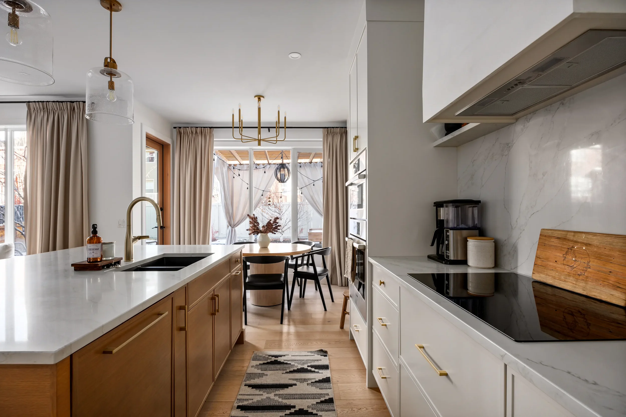 Interior view of a modern kitchen with white and wooden cabinetry, a white marble backsplash, a kitchen island with a white countertop, pendant lights, and large windows with beige curtains.