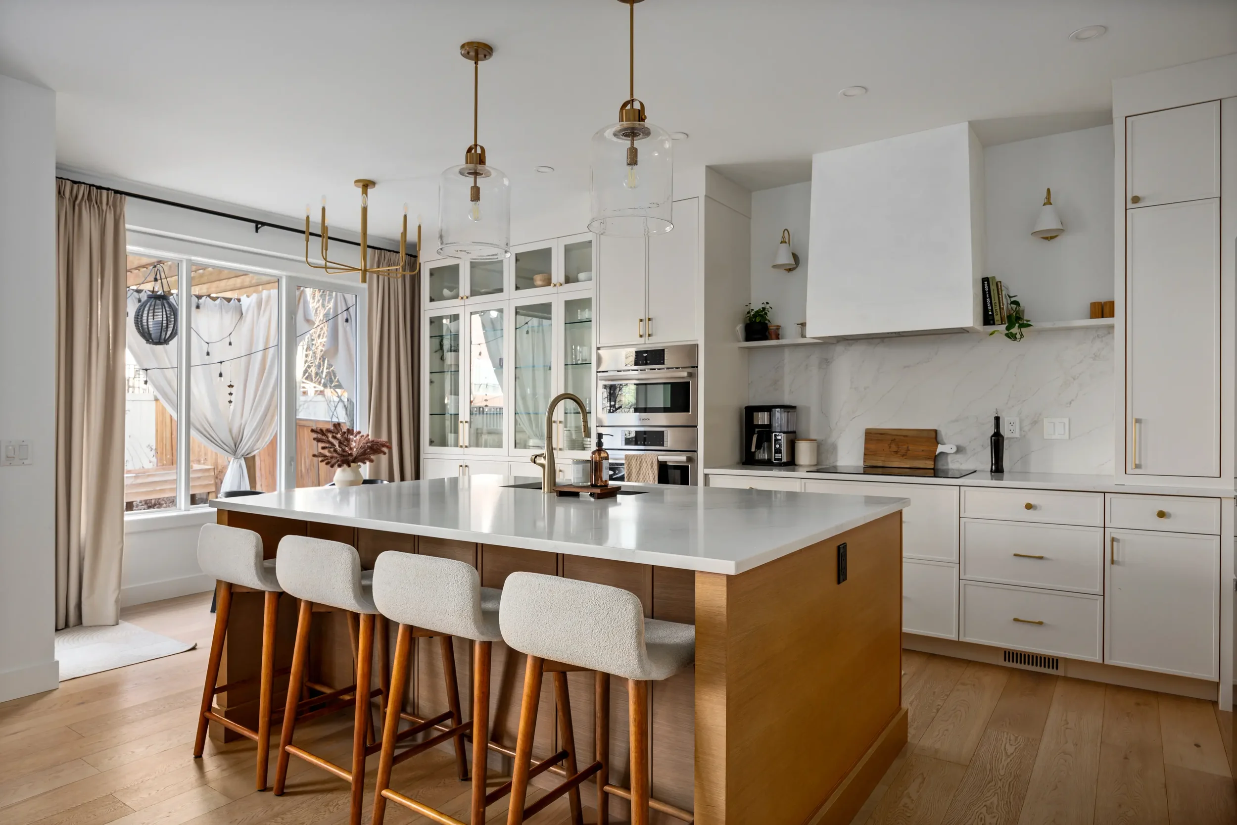 Modern kitchen with white cabinets, a center island with a white countertop, and pendant lights hanging above. Large window with beige curtains offers natural light. Wooden flooring and minimal decor with a few plants and books.