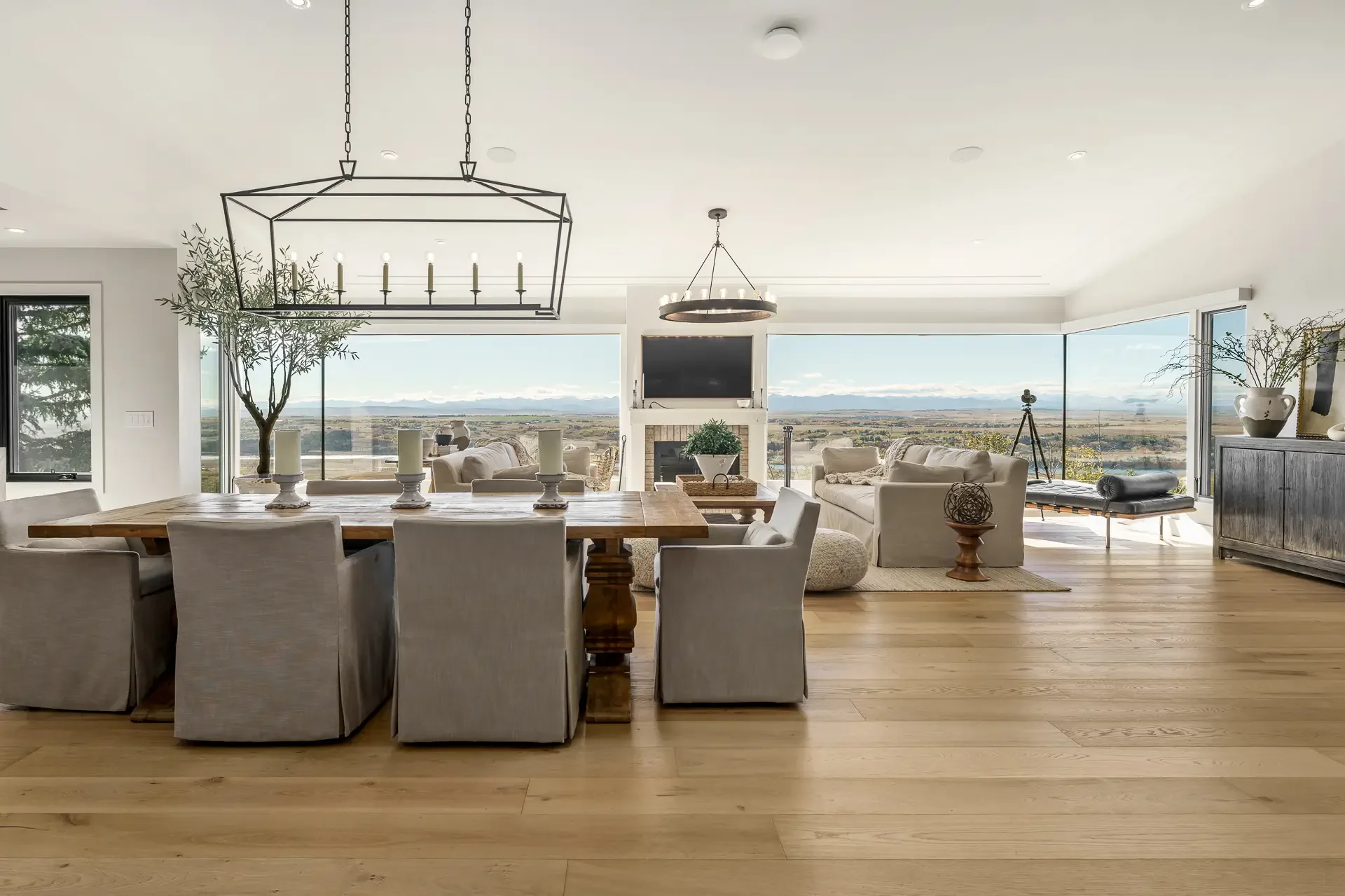 A spacious living room with large windows offering a view of open land and mountains. The room has white walls, wooden floors, a dining table with eight chairs, a sofa, a coffee table, plants, and modern lighting fixtures, including two geometric and