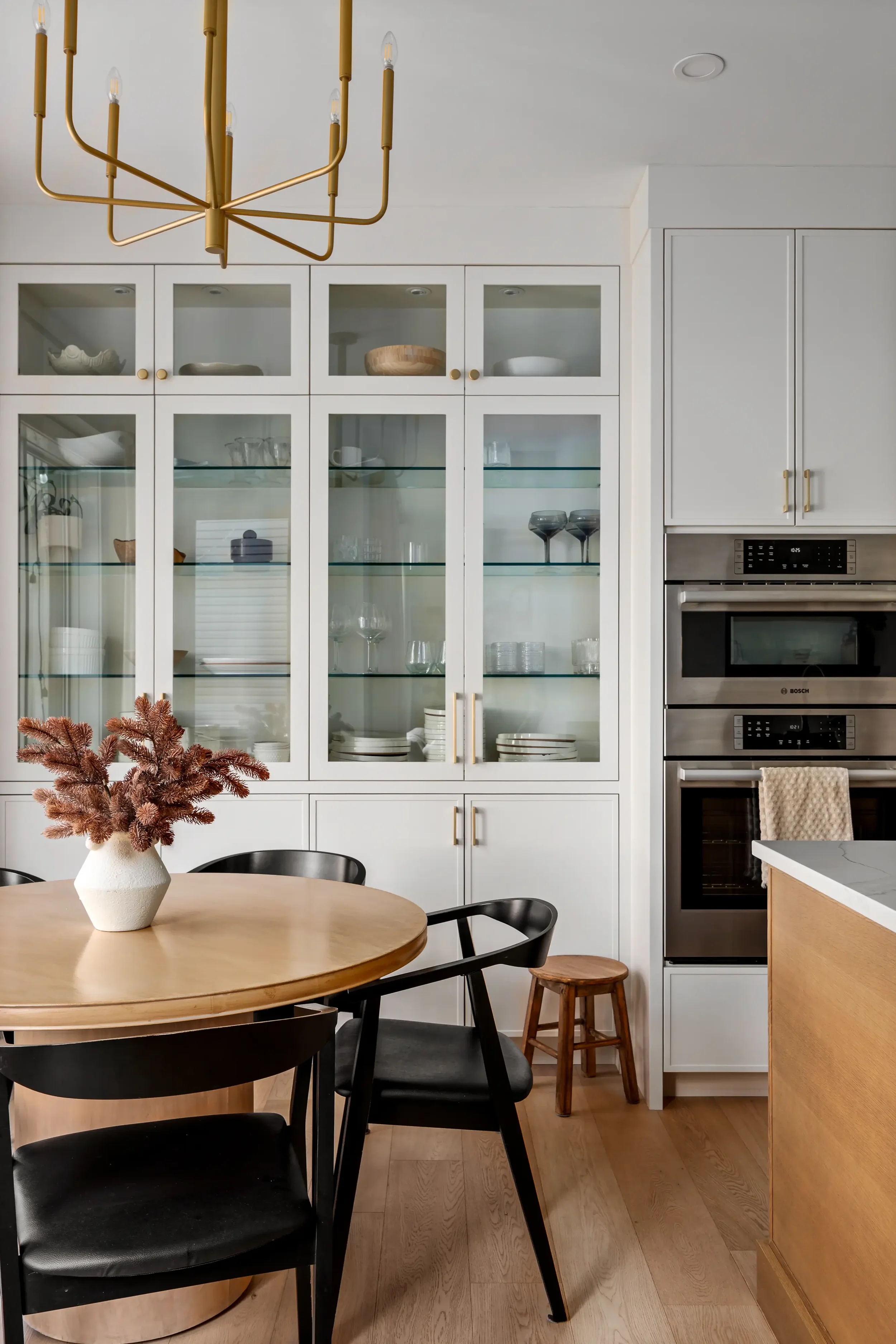 A kitchen with white cabinets, glass-front display cabinets, a built-in oven, a round wooden table with black chairs, a small wooden stool, a white vase with dried pinkish-brown flowers, and a gold chandelier hanging from the ceiling.