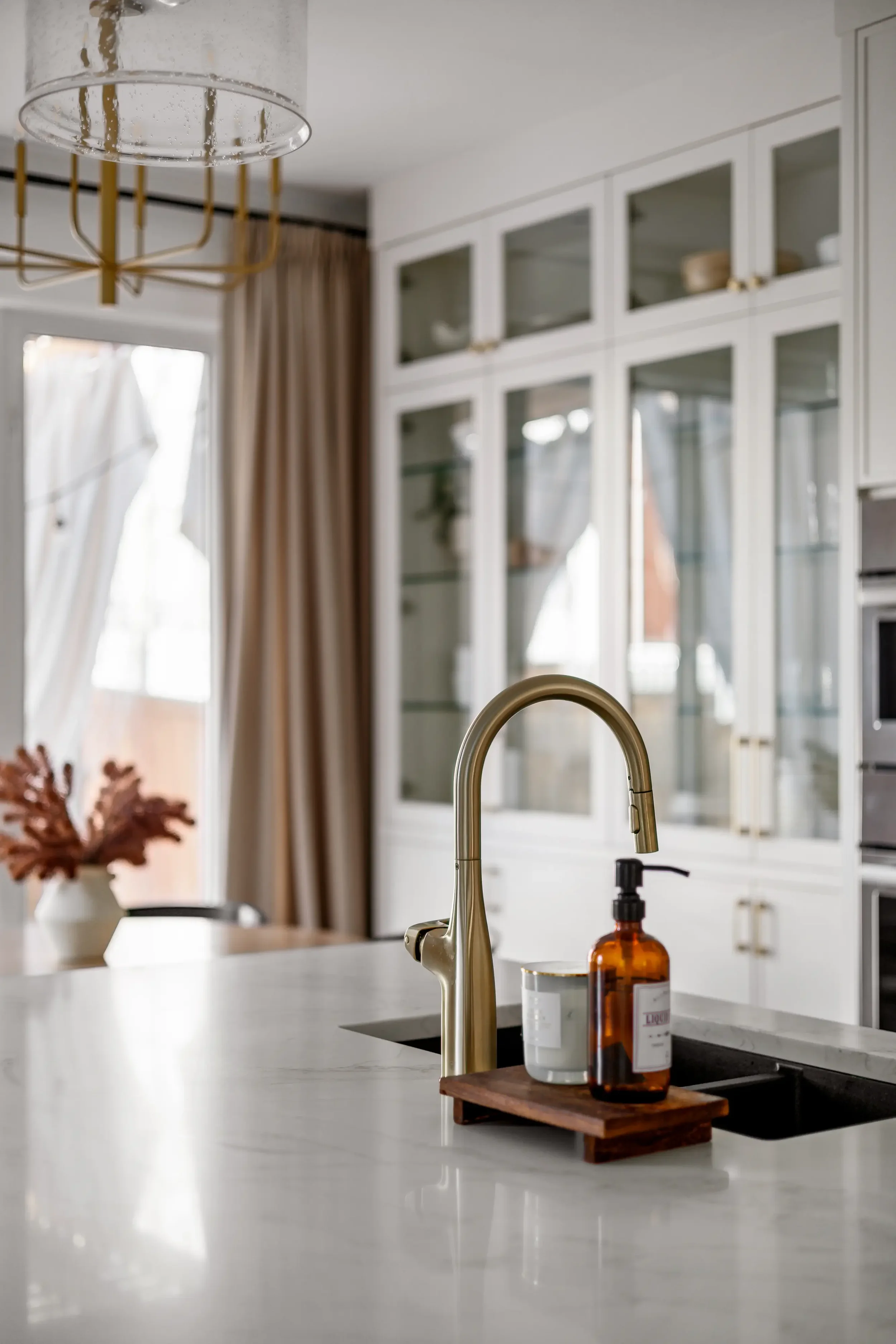 Close-up of a kitchen island with a gold faucet, a brown soap dispenser, and a small cutting board with bottles on it, in a modern kitchen with white cabinets and a window with curtains.