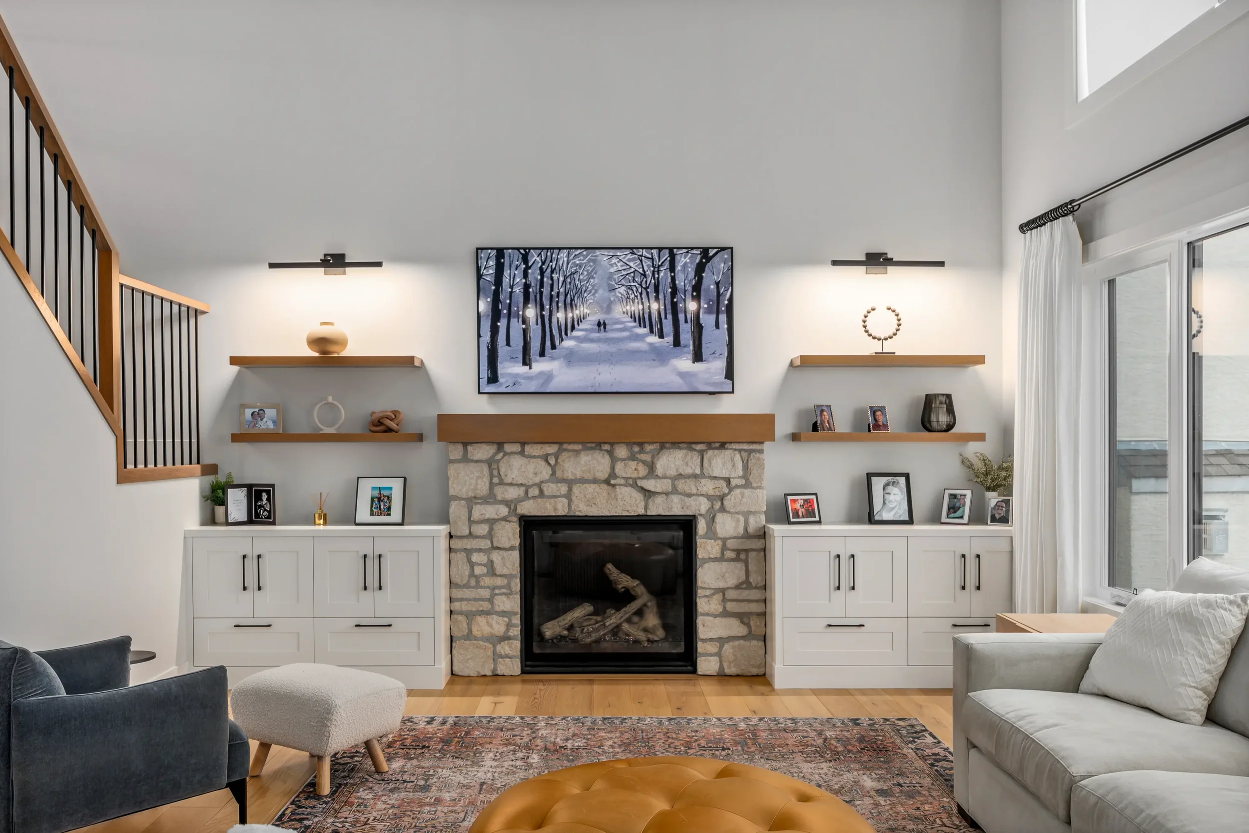 Living room with white cabinets, a stone fireplace, and floating shelves decorated with photos, vases, and artwork. A large window with curtains on the right lets in natural light. A ceiling-mounted TV displays a snowy winter scene with trees and a pathway. Furniture includes a black chair, white ottoman, and a beige sofa, with a patterned rug and a tufted leather ottoman.