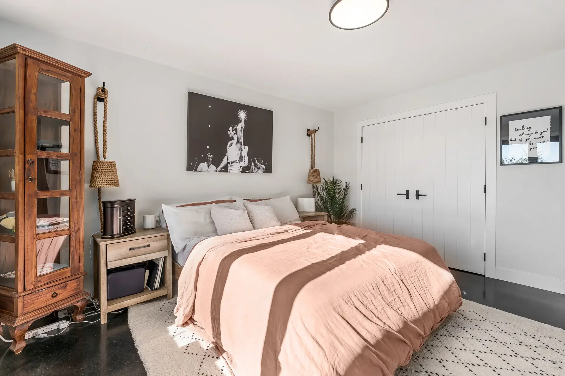 A cozy bedroom with a bed covered in a pink and brown striped comforter, two white pillows, and a small bedside table with a lamp and decorative accents. On the left, a large glass-front wooden cabinet holds various items, and a rope hanging on the w