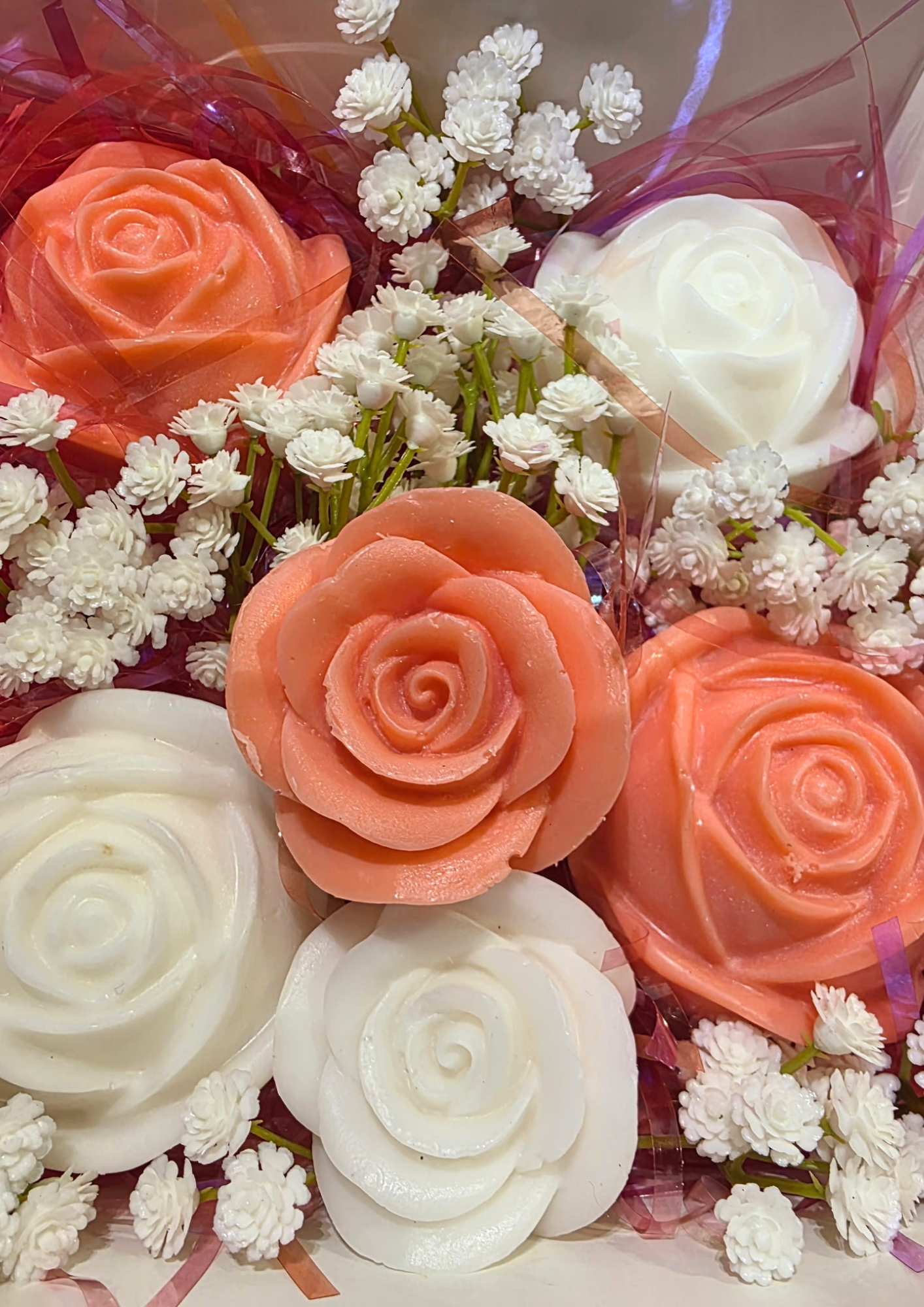 Bouquet of rose-shaped soaps in white, peach, and pink, surrounded by small white flowers and wrapped in decorative pink and purple cellophane.