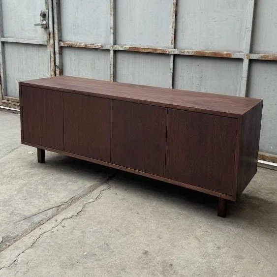 A long, dark wood sideboard with four cabinet doors, standing on four legs, against a garage door or metal wall in a concrete floor setting.