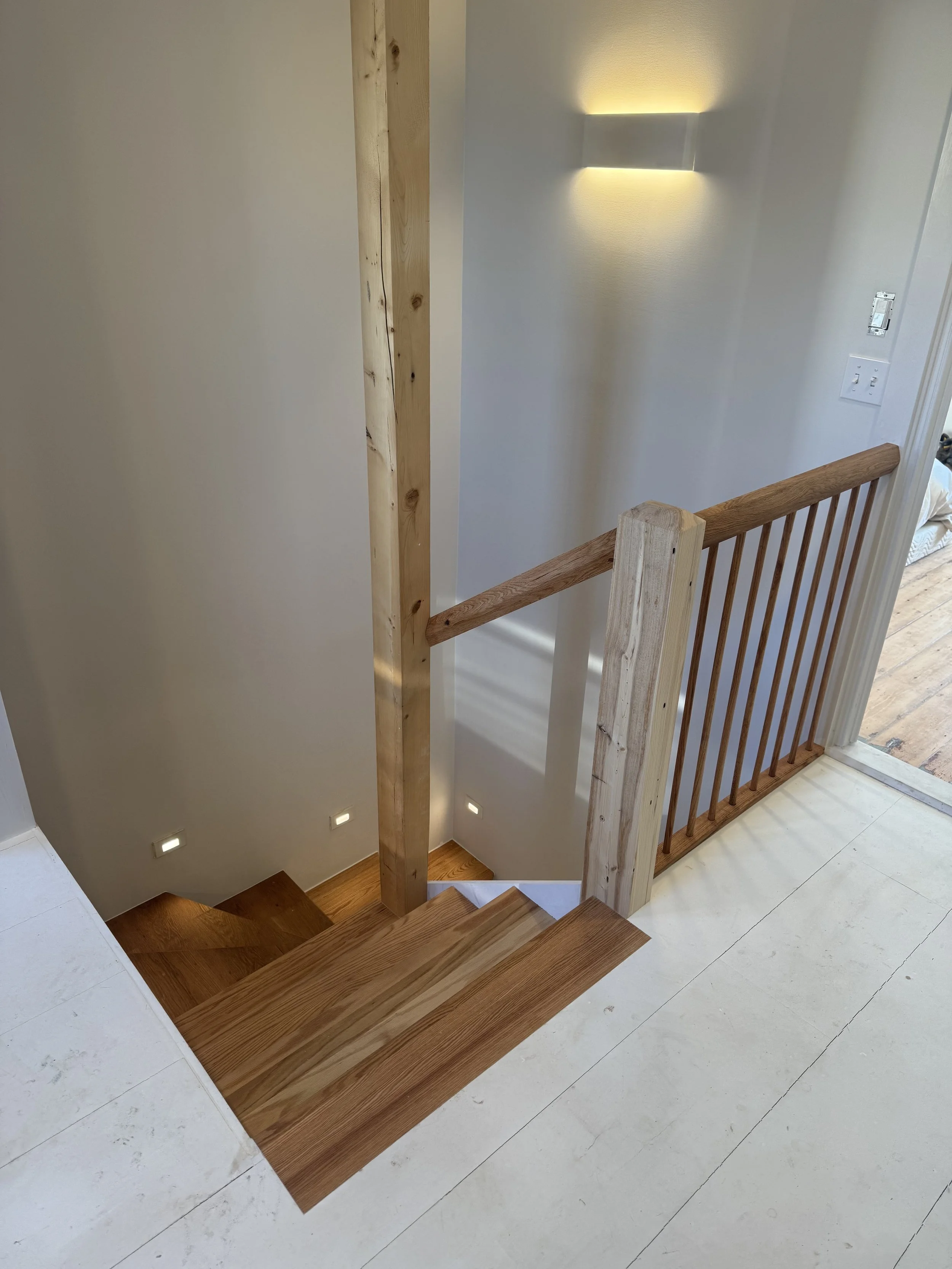 Photo of a staircase with wooden steps, handrail, and support post inside a home with white walls and minimal lighting.