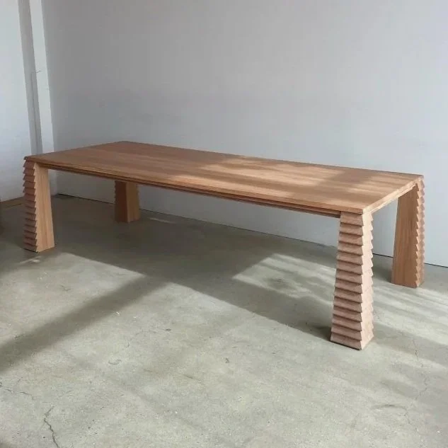 A wooden table with a smooth top and side legs made of stacked rectangular blocks, placed on a concrete floor in a minimalist room.