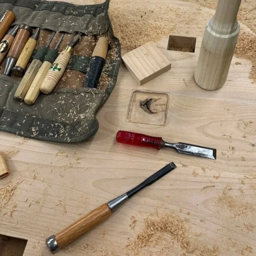 Work tools on a woodworking bench, including chisels, a chisel, a wood piece, a hammer, and a carving gouge.