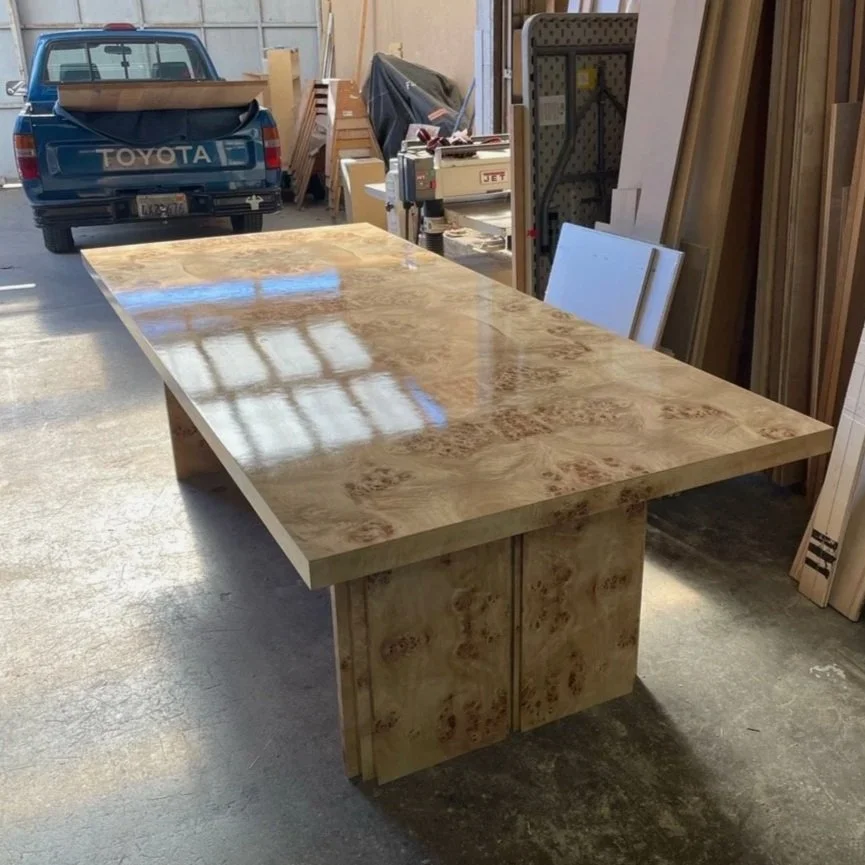 A large wooden table with a glossy finish in a woodworking shop, with wood materials and tools in the background.