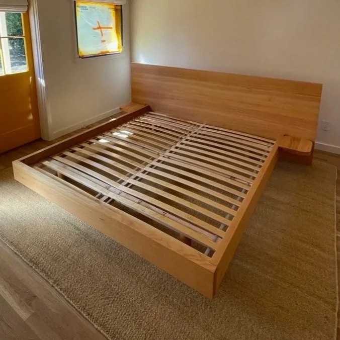 A wooden bed frame without a mattress in a bedroom with a window and a door, sunlight casting shadows on the floor.