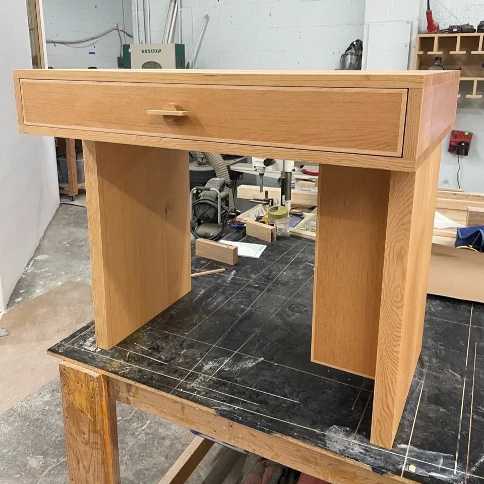 A wooden piece of furniture, possibly a desk or console table, being built in a workshop. It has a long drawer on top with a small wooden handle, standing on two vertical panels.