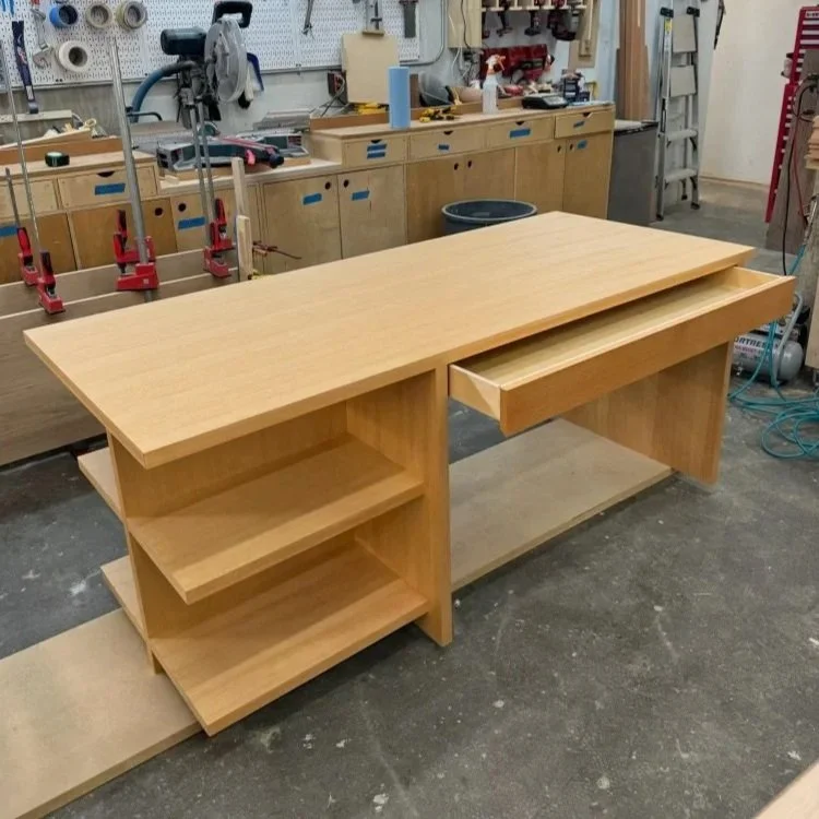 Wooden desk with open shelves on the left side and a pull-out keyboard drawer on the right side, in a woodworking workshop.