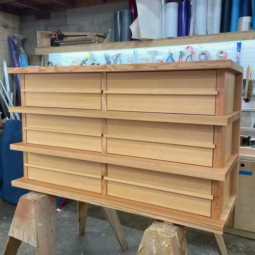 A wooden dresser with multiple drawers in a woodworking shop, with tools and supplies on a pegboard and shelves in the background.