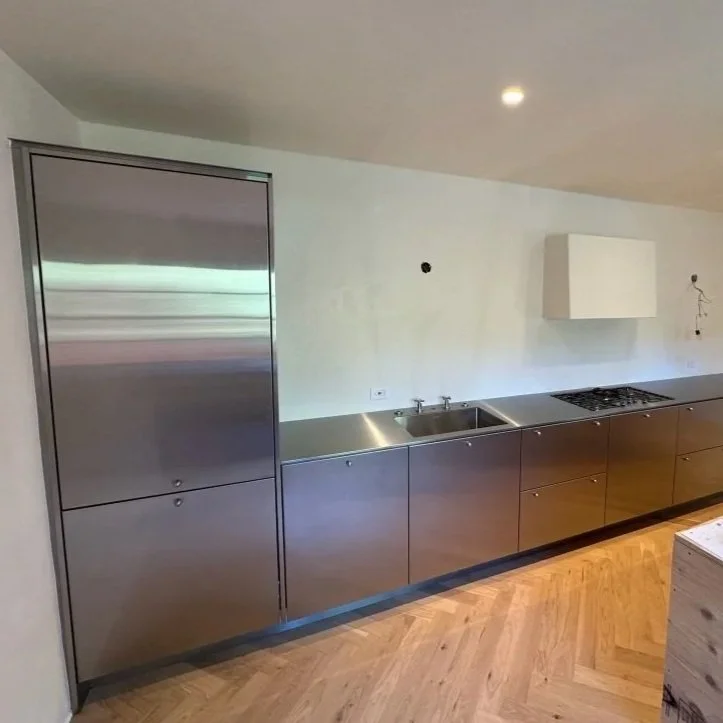 Modern kitchen with stainless steel cabinets, a built-in refrigerator, a sink, a stove, and a white wall-mounted cabinet, with wooden flooring.