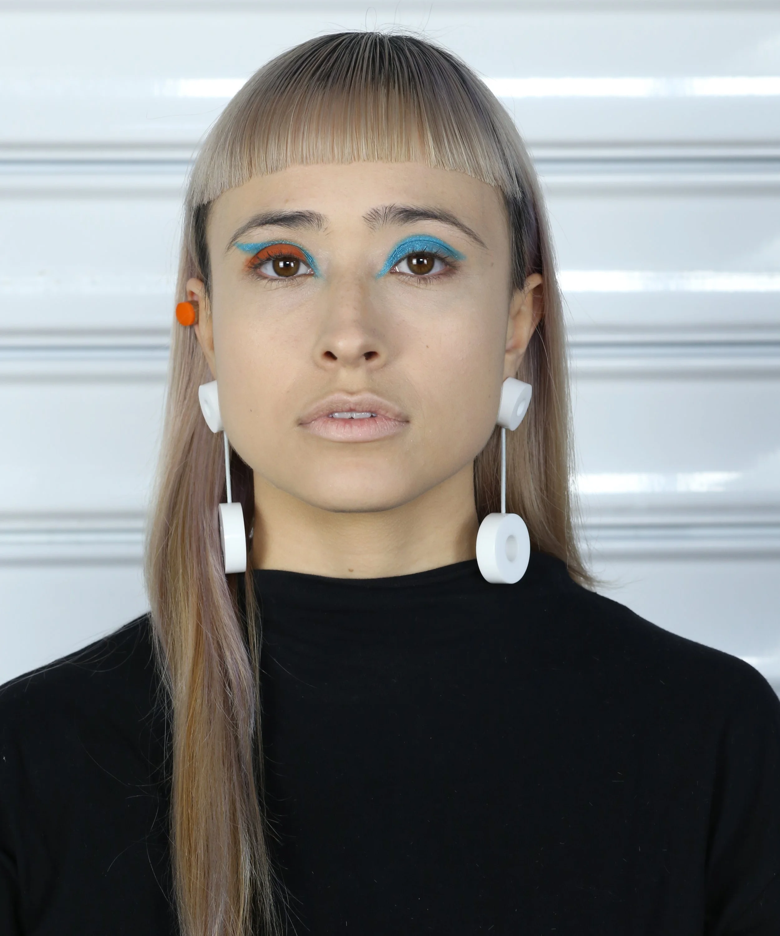 Close-up of a woman with multicolored eye makeup and long hair, wearing white wireless earbuds against a metallic background.