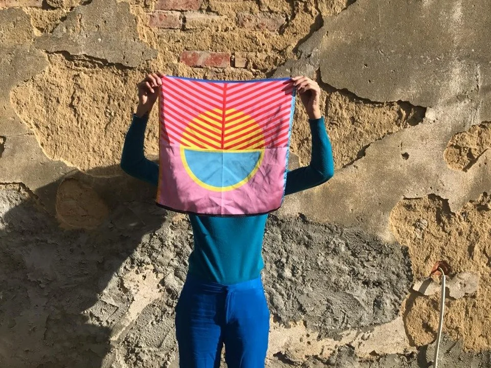 Person standing against a stone wall holding a colorful flag in front of their face, obscuring their identity.