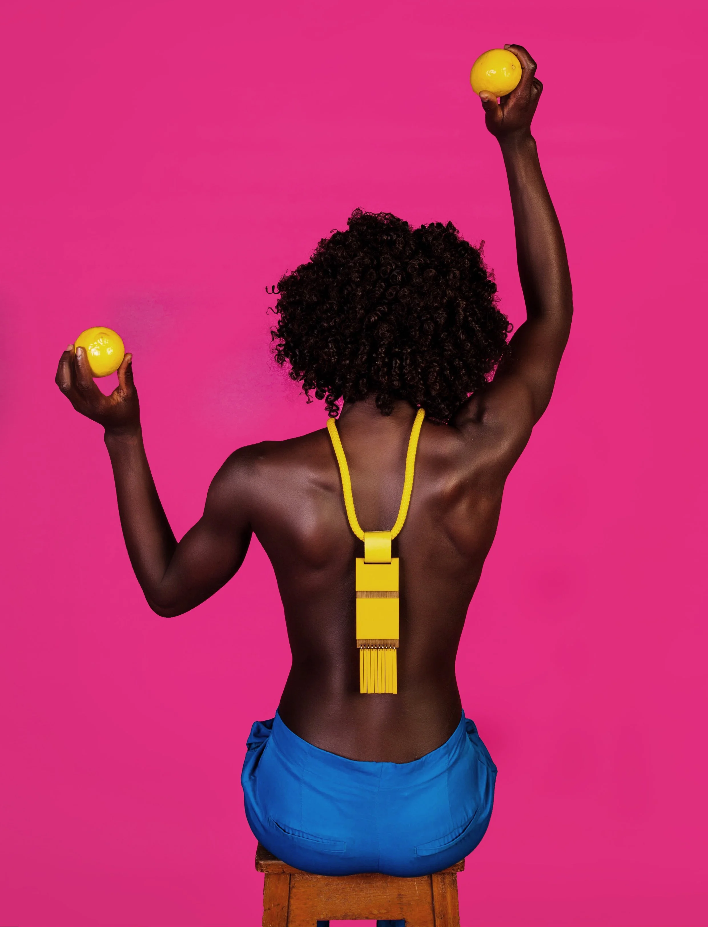Back view of a person with dark skin and curly hair sitting on a wooden stool, holding yellow objects in each hand, wearing blue shorts, a yellow necklace, and a yellow pendant against a pink background.