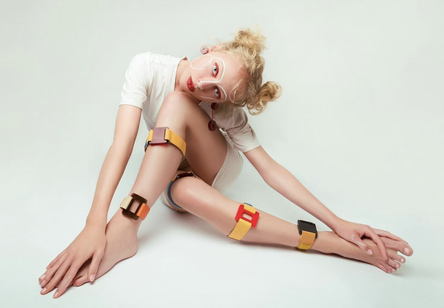 A woman with curly blonde hair in a stretch pose on the floor, wearing a white t-shirt, with colorful plastic clips attached to her arms, legs, and neck, and a transparent face shield.