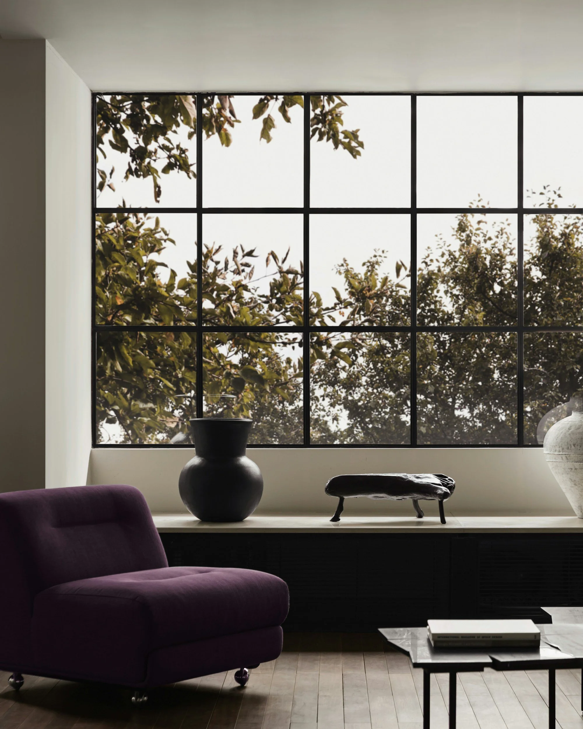 Interior room with a purple armchair, large window with black grid, green trees outside, and decorative objects on a ledge including a black vase, a small black sculpture, and a white vase.