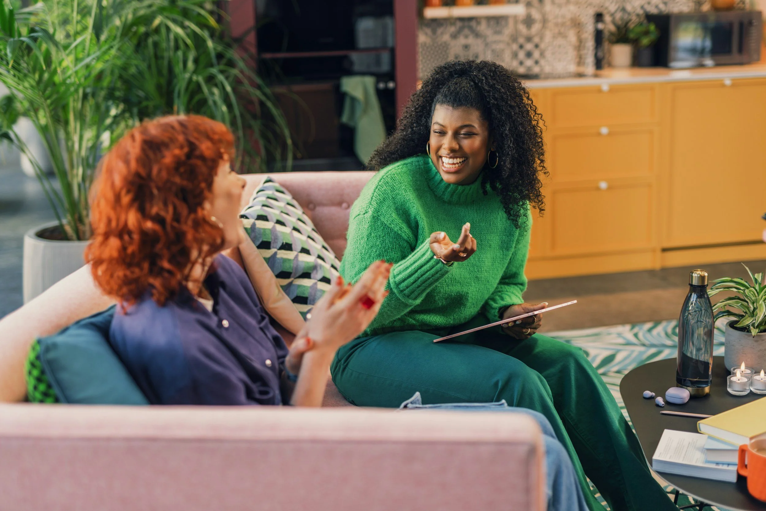 Two women sitting on a pink couch in a cozy living room, talking happily. One woman has curly black hair and wears a green sweater, the other has straight red hair and wears a purple shirt. There is a coffee table with books, candles, a bottle, and a small plant in front of them.