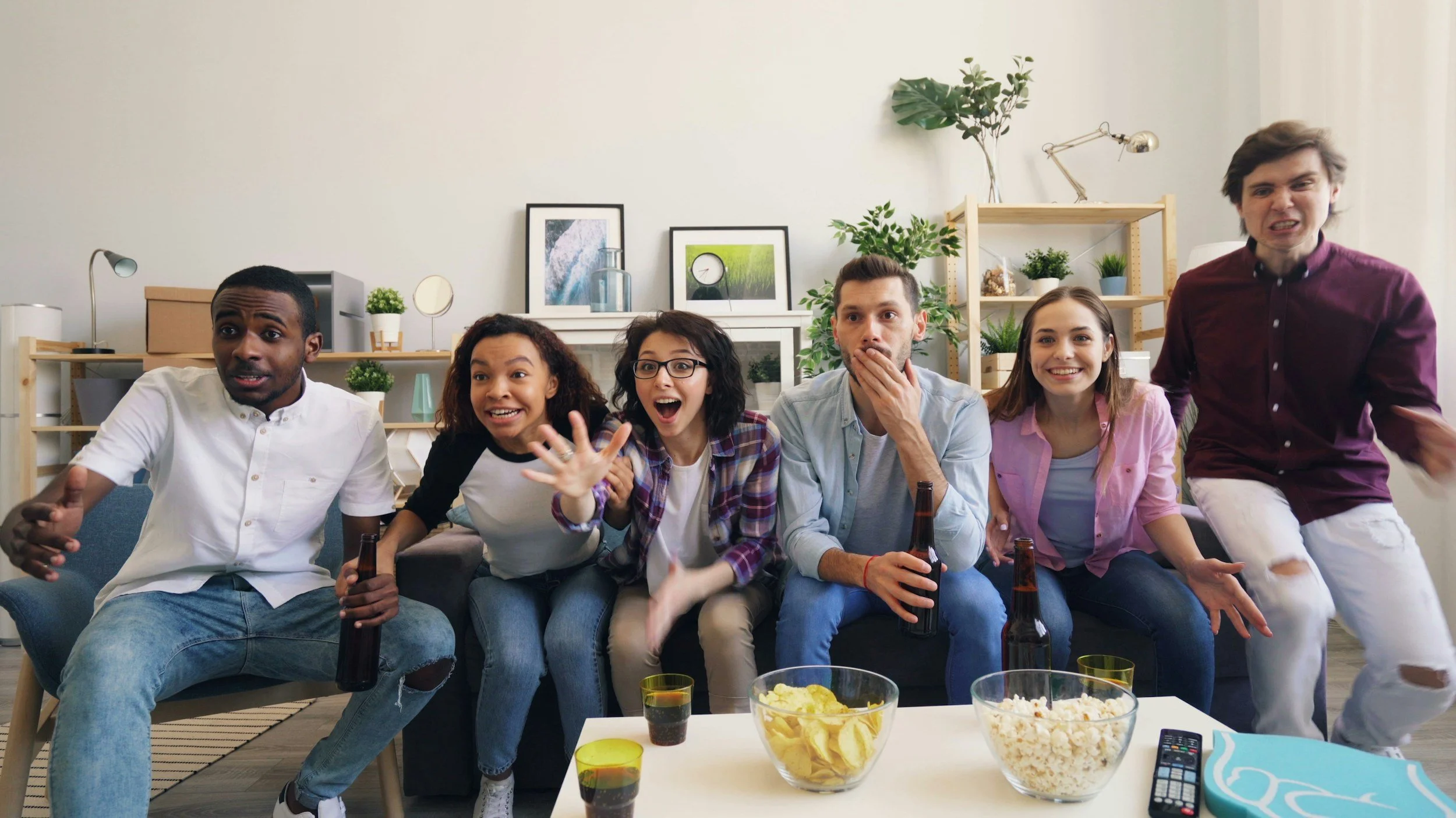 A diverse group of six young adults sitting on a couch, reacting excitedly to a game or event on TV, with snacks and drinks on the table in front of them.