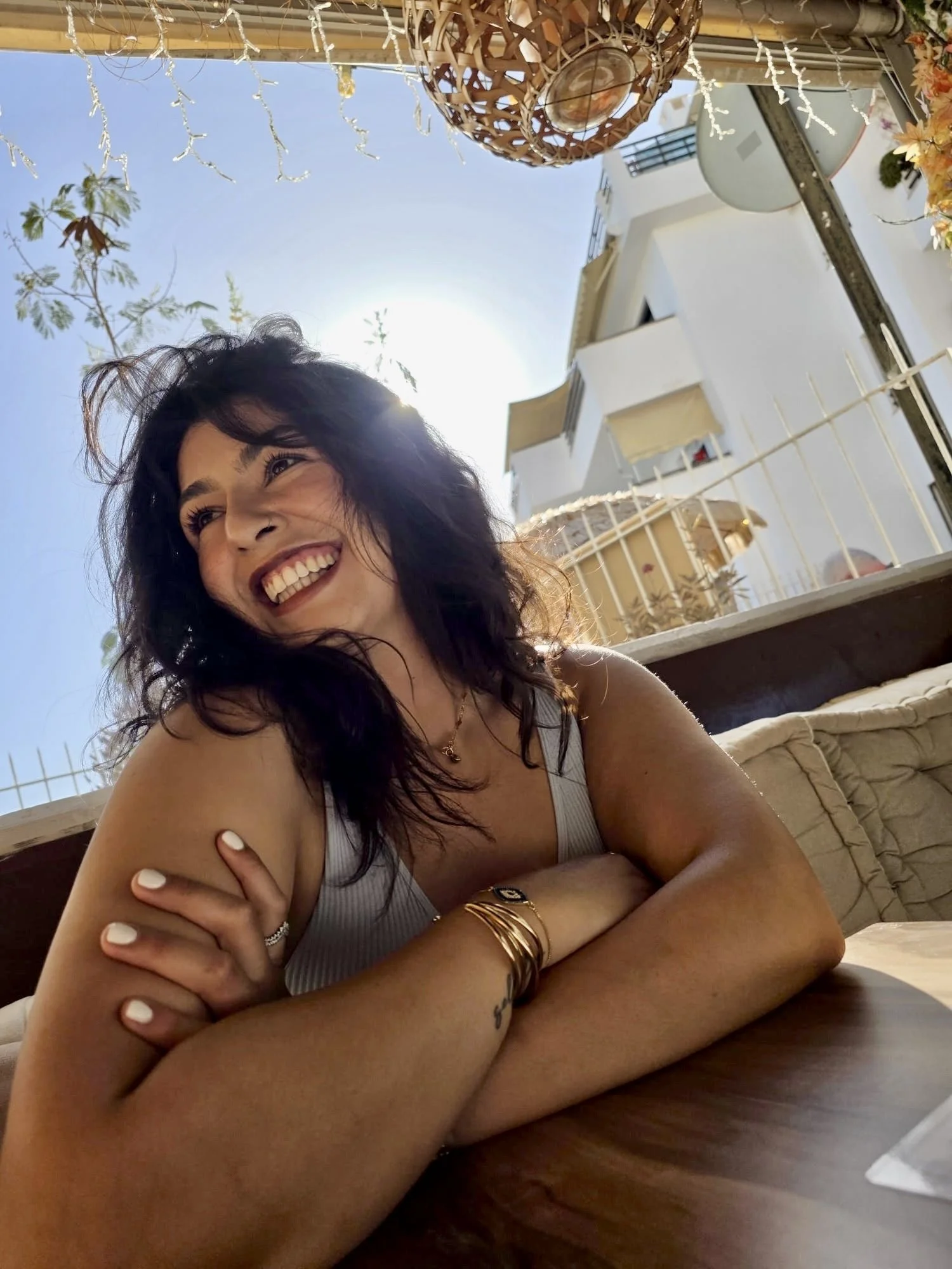 A young woman with dark curly hair smiling at a cafe, sunlight shining behind her, with outdoor buildings and blue sky in the background.