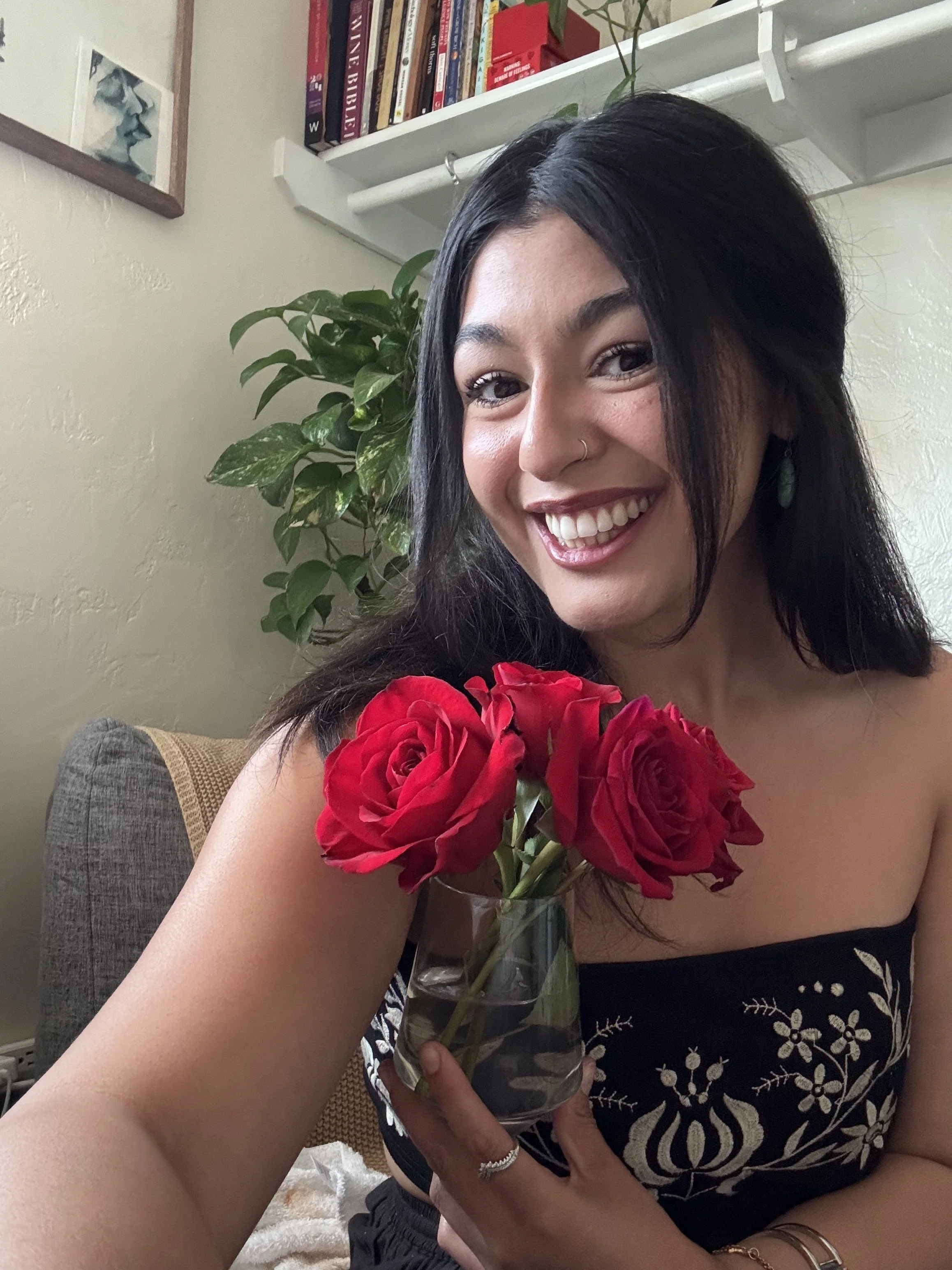 A woman with dark hair and a nose ring smiling while holding a glass vase with red roses, sitting on a couch with a beige cushion in a room with bookshelves, a potted plant, and framed artwork in the background.