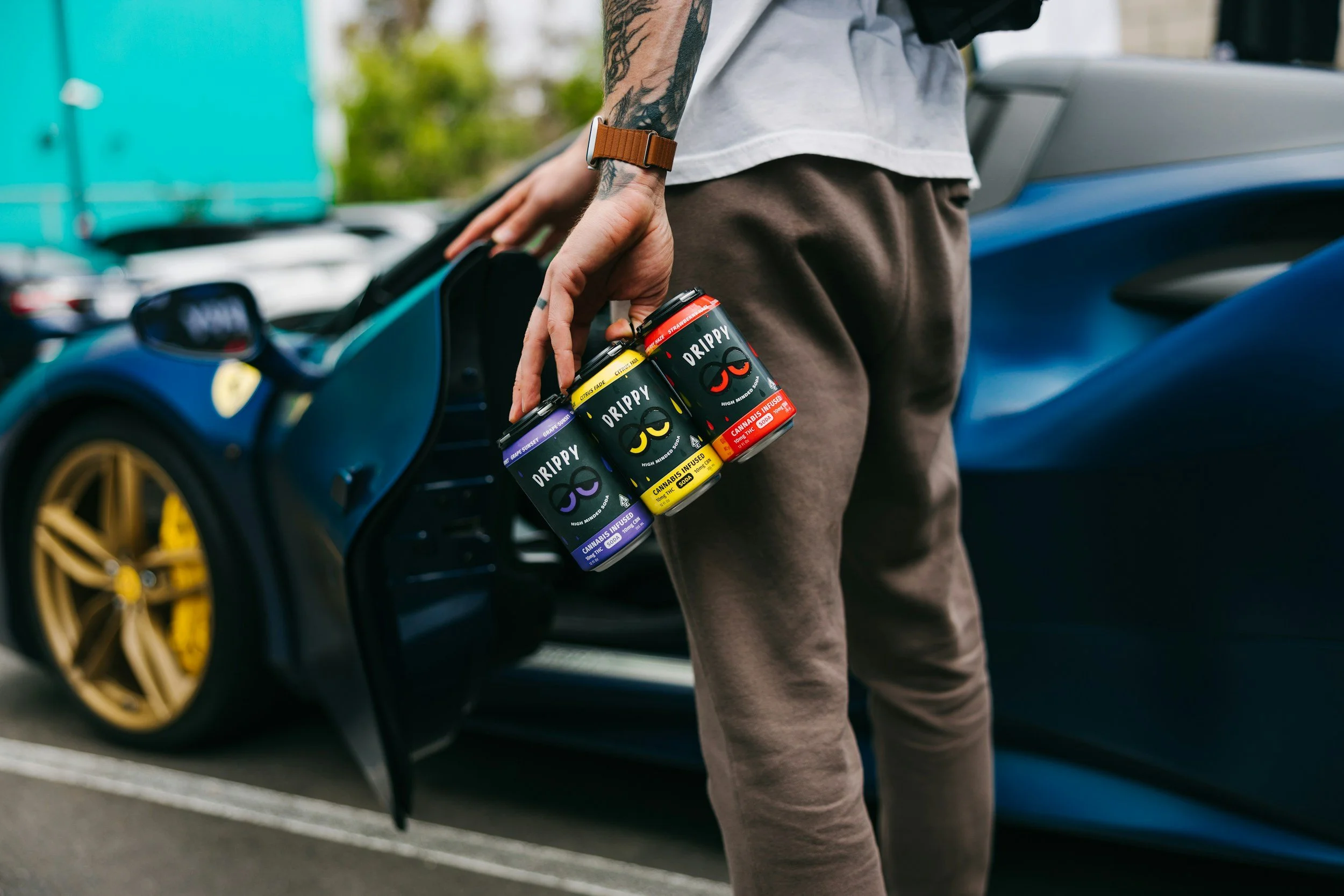 Person holding four cans of cannabis-infused beverage standing next to a blue sports car with gold wheels.