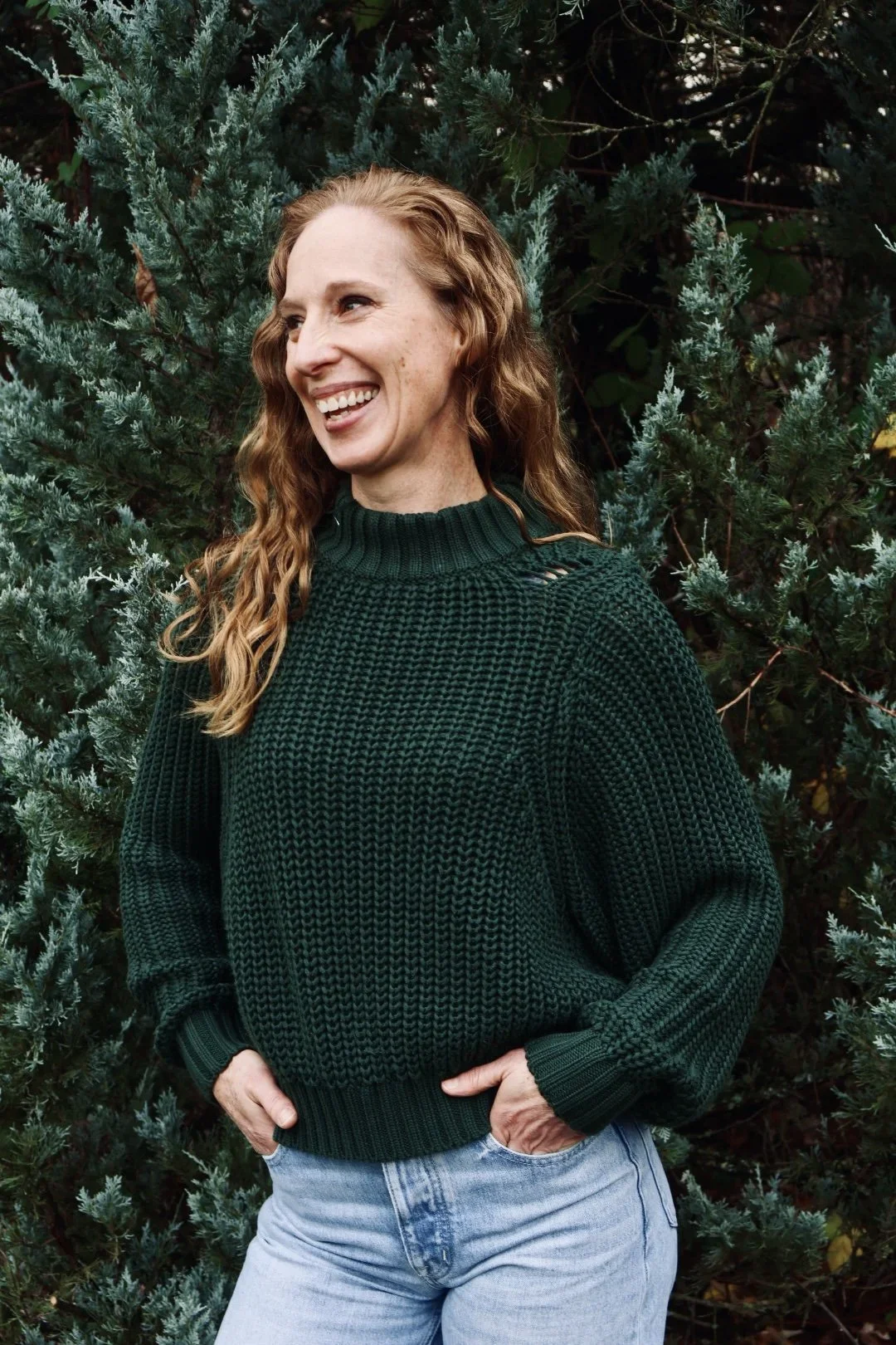 A woman with curly, red hair, wearing a dark green knitted sweater and light blue jeans, smiling while standing in front of dense greenery.