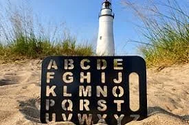 A black stencil letterboard placed on a sandy area with grass and a lighthouse in the background.