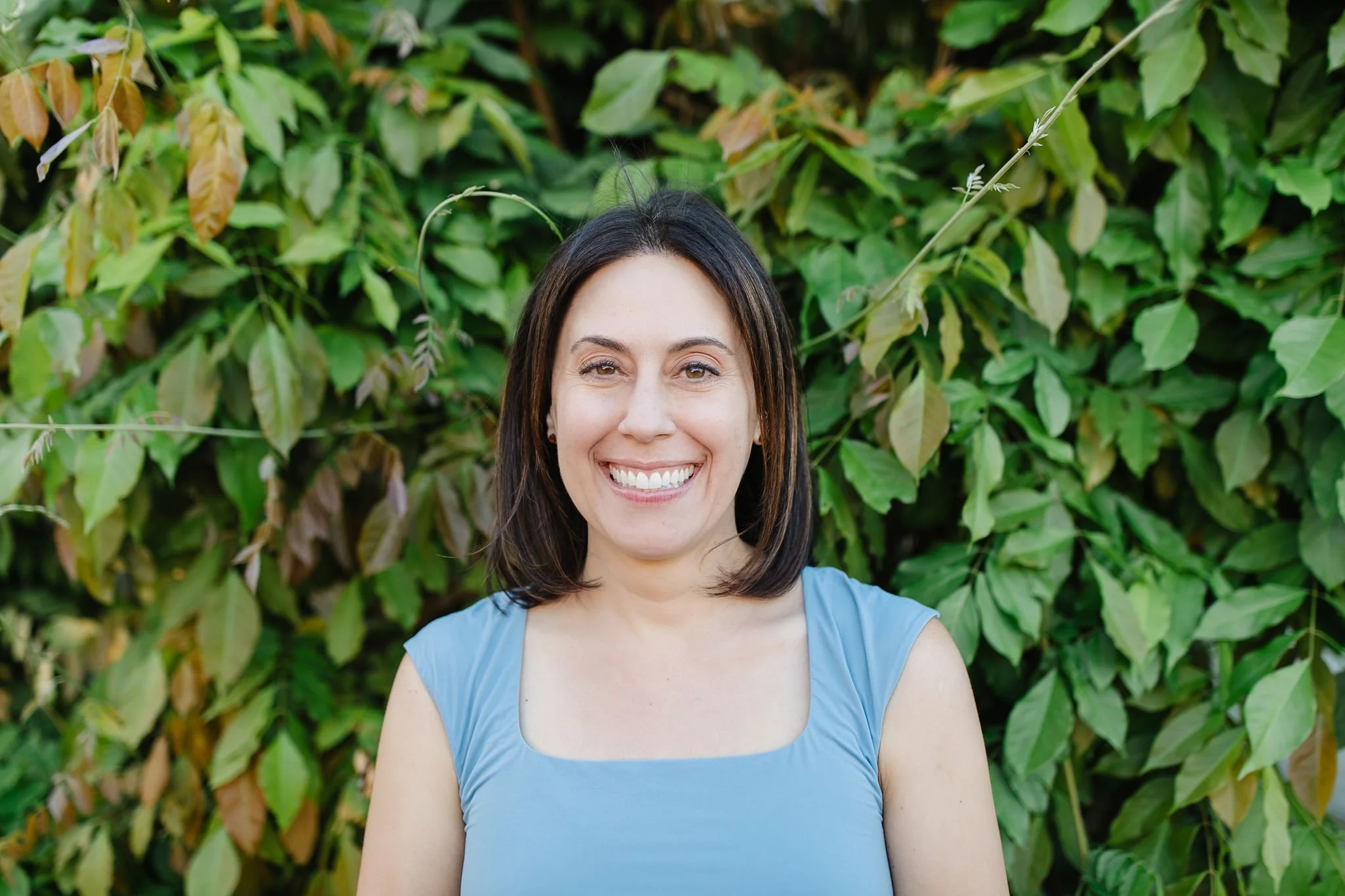 Woman with shoulder-length dark hair smiling in front of green leafy bushes, wearing a sleeveless light blue top.
