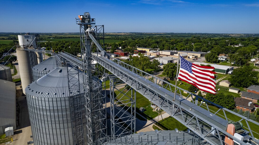 An aerial view of a grain silo facility with an American flag, surrounded by green fields and a small town.