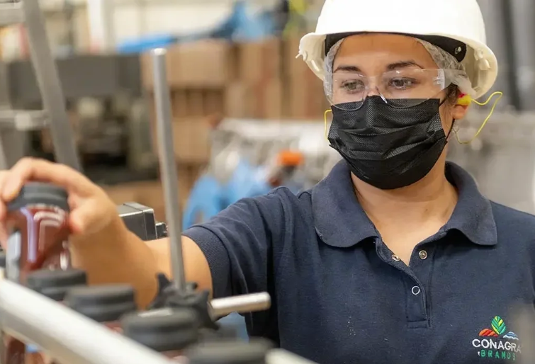 A woman working in a manufacturing or industrial setting, wearing a white safety helmet, clear safety glasses, a black face mask, and a navy blue polo shirt with a logo that reads 'Conagra Brands.'