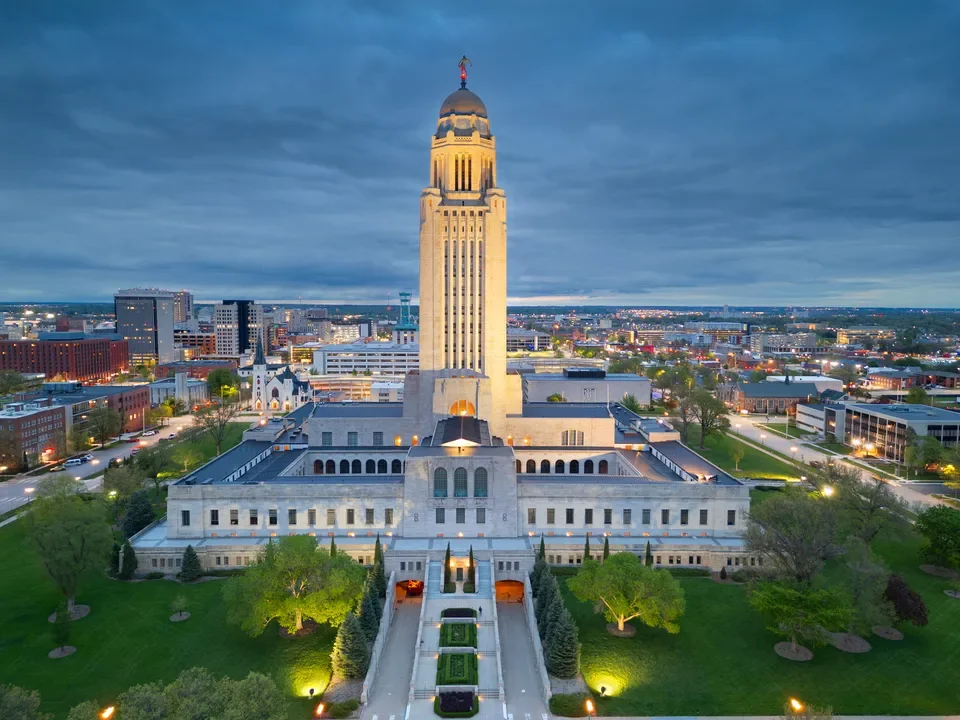 Nighttime view of the Nebraska State Capitol building in Lincoln, Nebraska, illuminated with city lights in the background.