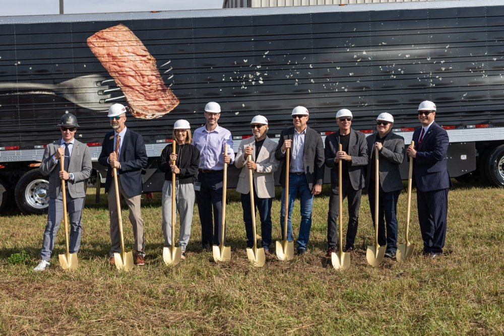 Group of nine suited men and women wearing construction helmets, holding shovels, at a groundbreaking event in front of a truck with an image of a steak on it.