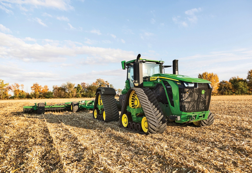 Green John Deere tractor with wide tracks pulling a large farming implement across a harvested field under a blue sky with clouds.