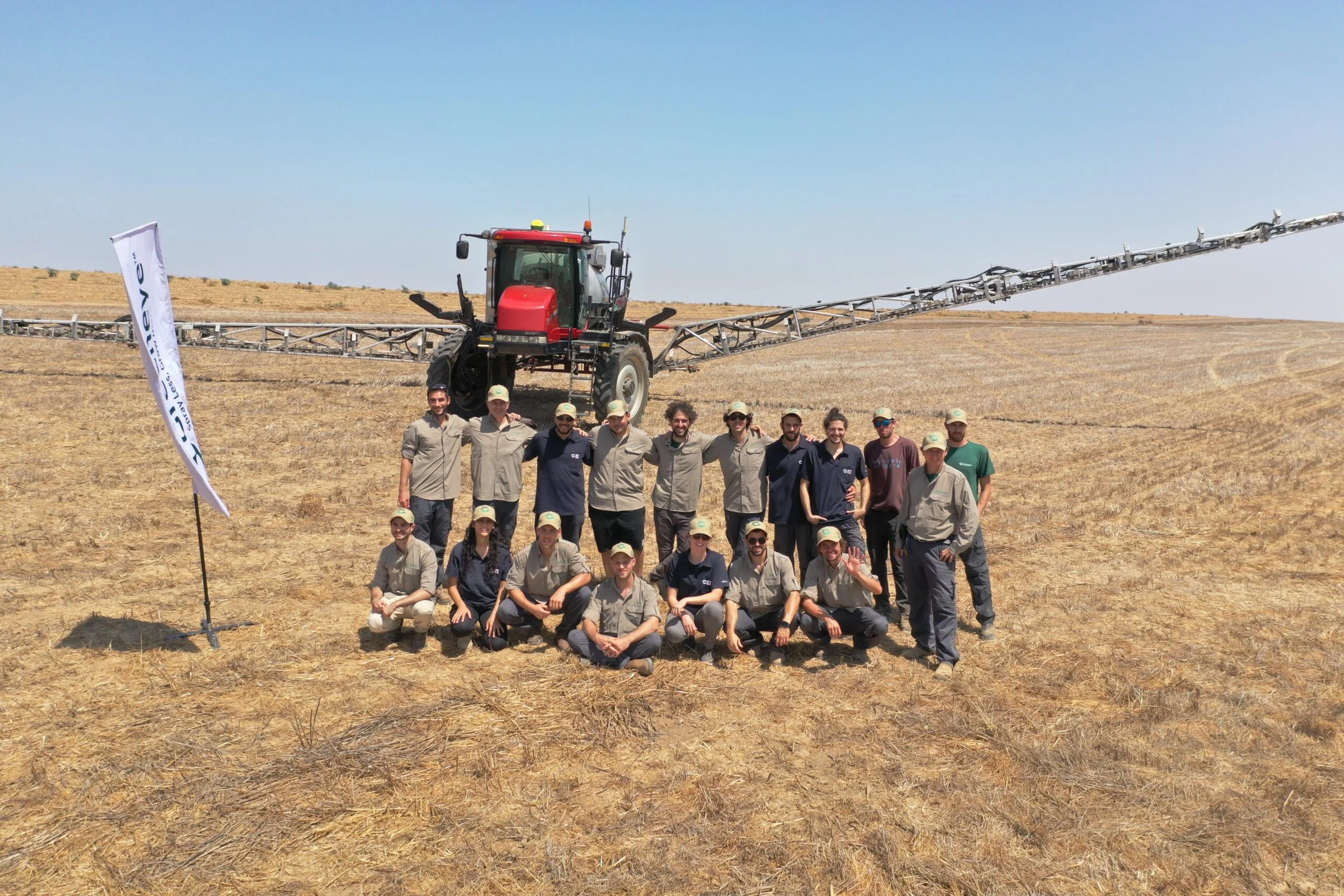 A group of people standing in front of a large red agricultural sprayer machine in a dry field under a clear blue sky.