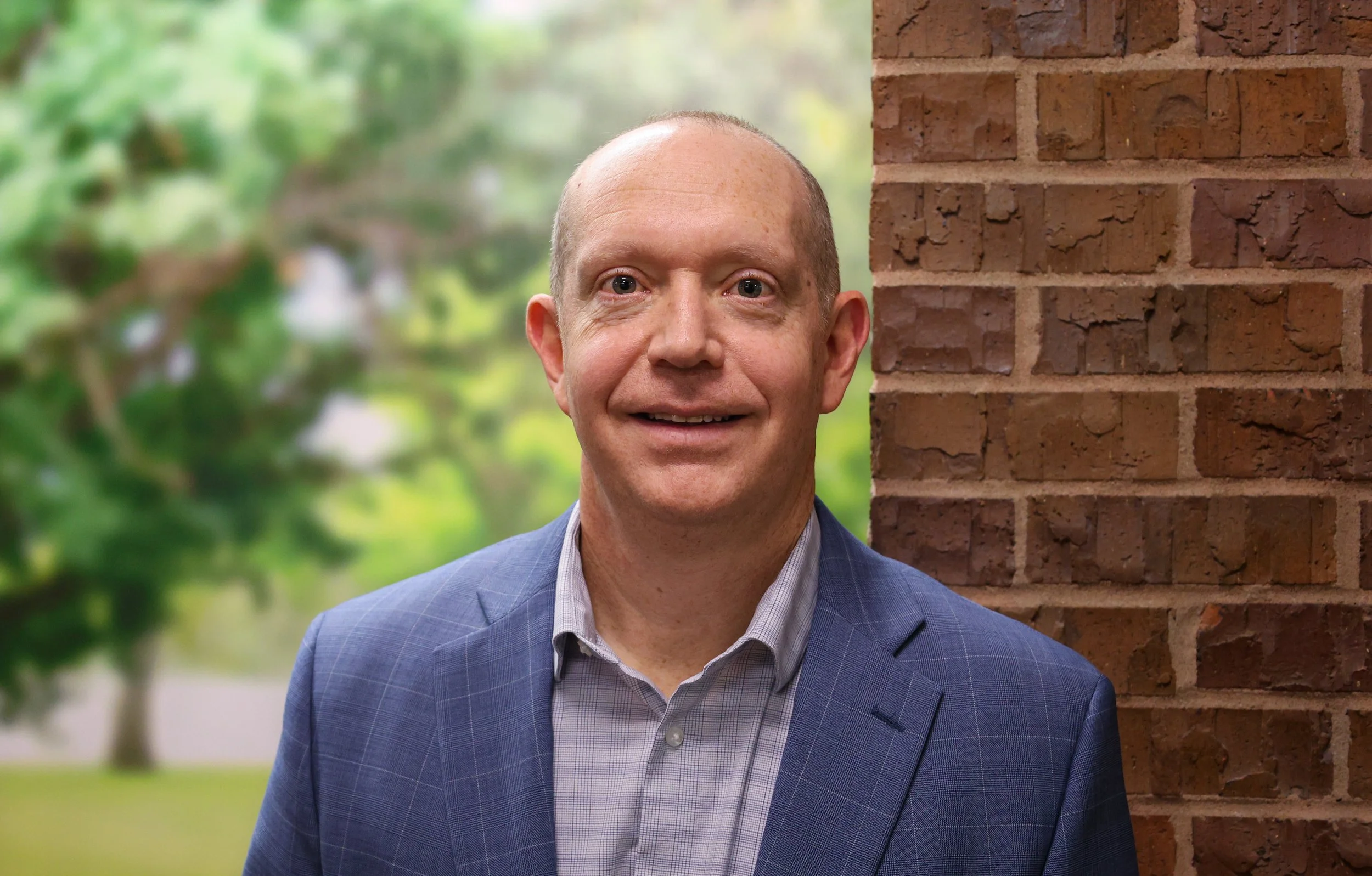 A middle-aged man with a receding hairline, wearing a blue checked blazer and a white shirt, standing next to a brick wall with a blurred green outdoor background.