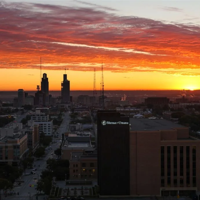 City skyline at sunset with a vibrant orange and pink sky, tall buildings, and radio towers.