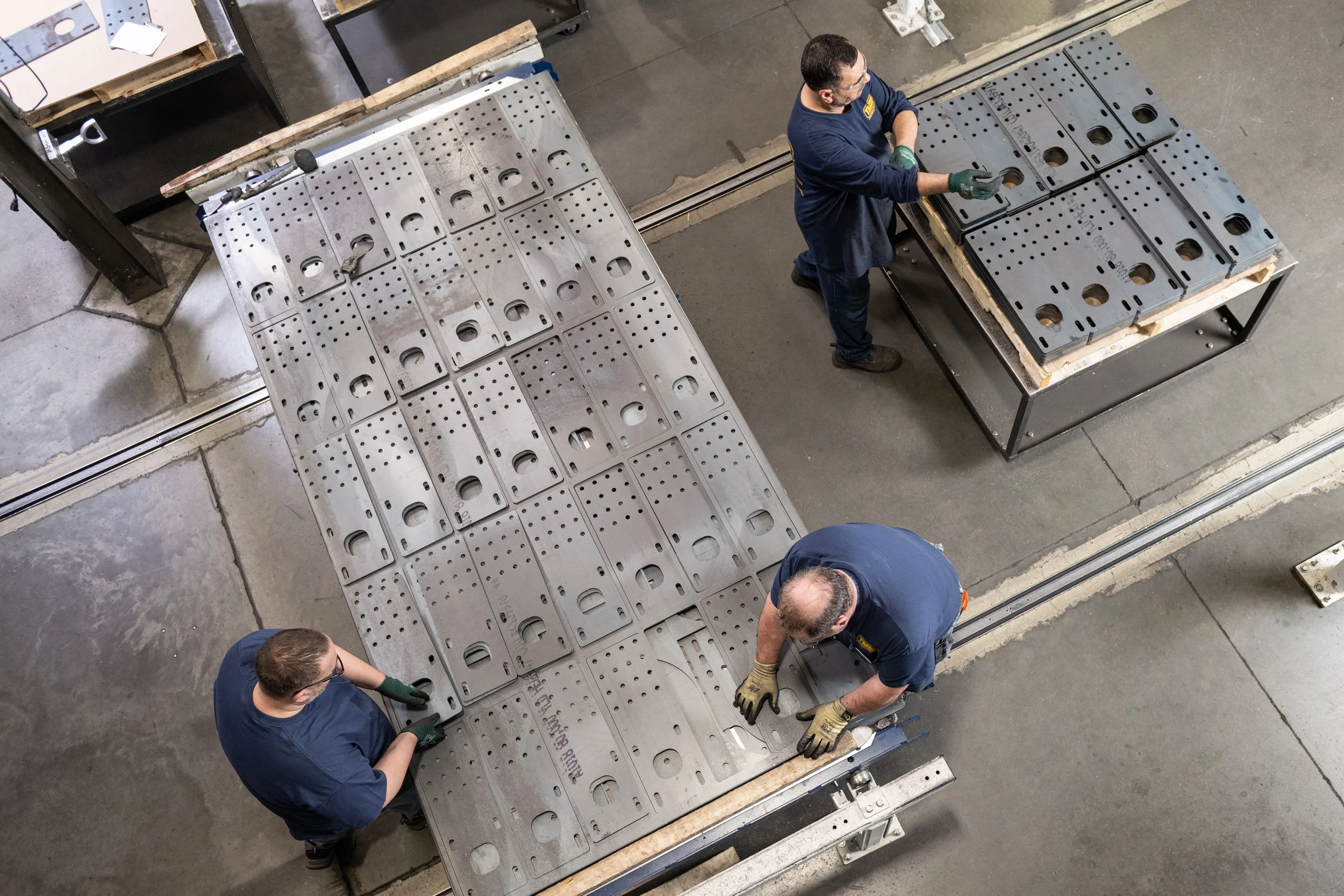 Workers assembling or inspecting large metal plates with holes on a worktable in an industrial setting.