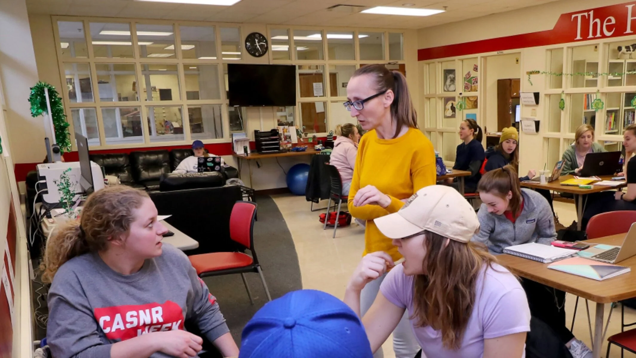 A group of students and a teacher in a classroom engaged in conversation. Students are seated at tables with laptops and notebooks, and the teacher stands nearby. The classroom has a red and beige color scheme, a wall clock, and a glass window partition.