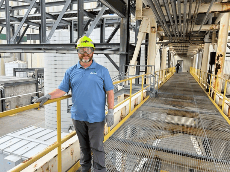 A man wearing a Cargill uniform, safety glasses, a yellow hard hat, and gloves standing on an industrial walkway with piping and machinery in the background.