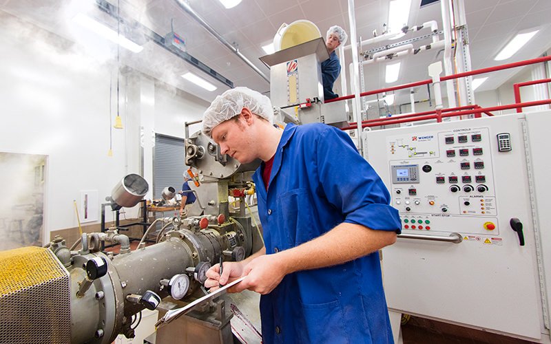 Two workers in a laboratory or industrial facility, wearing blue coveralls, hairnets, and gloves. One worker is operating a large scientific or industrial machine, taking notes on a clipboard. The other worker is standing on a platform in the background, observing or adjusting equipment. The environment is clean and equipped with control panels and gauges.
