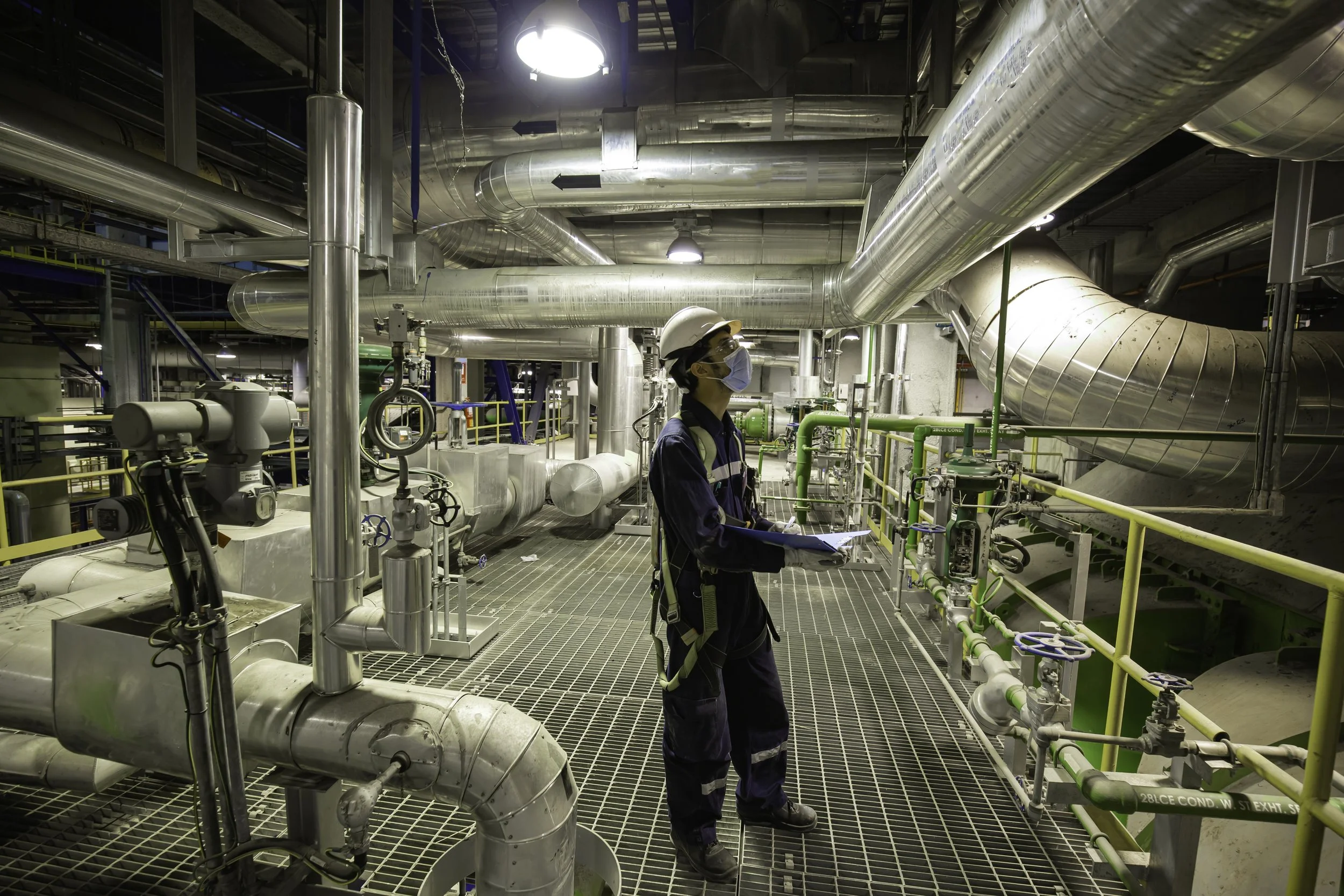 A person wearing safety gear, including a helmet, mask, and safety vest, inspecting industrial pipes and equipment in a large facility with metal pipes, gauges, and ventilation ducts.