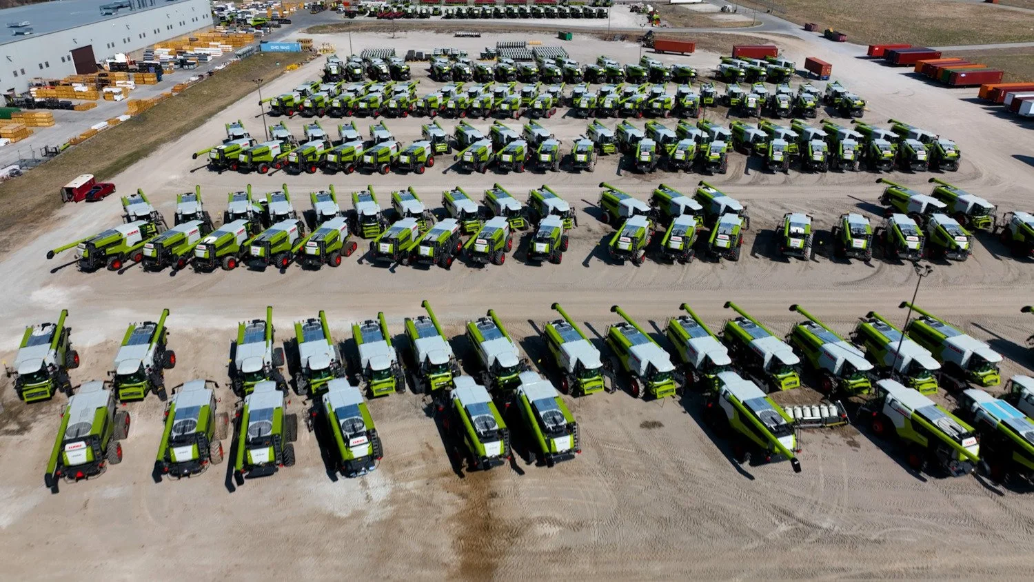 Aerial view of an outdoor lot filled with numerous green and white street sweepers parked in rows, with a large industrial building and other vehicles in the background.