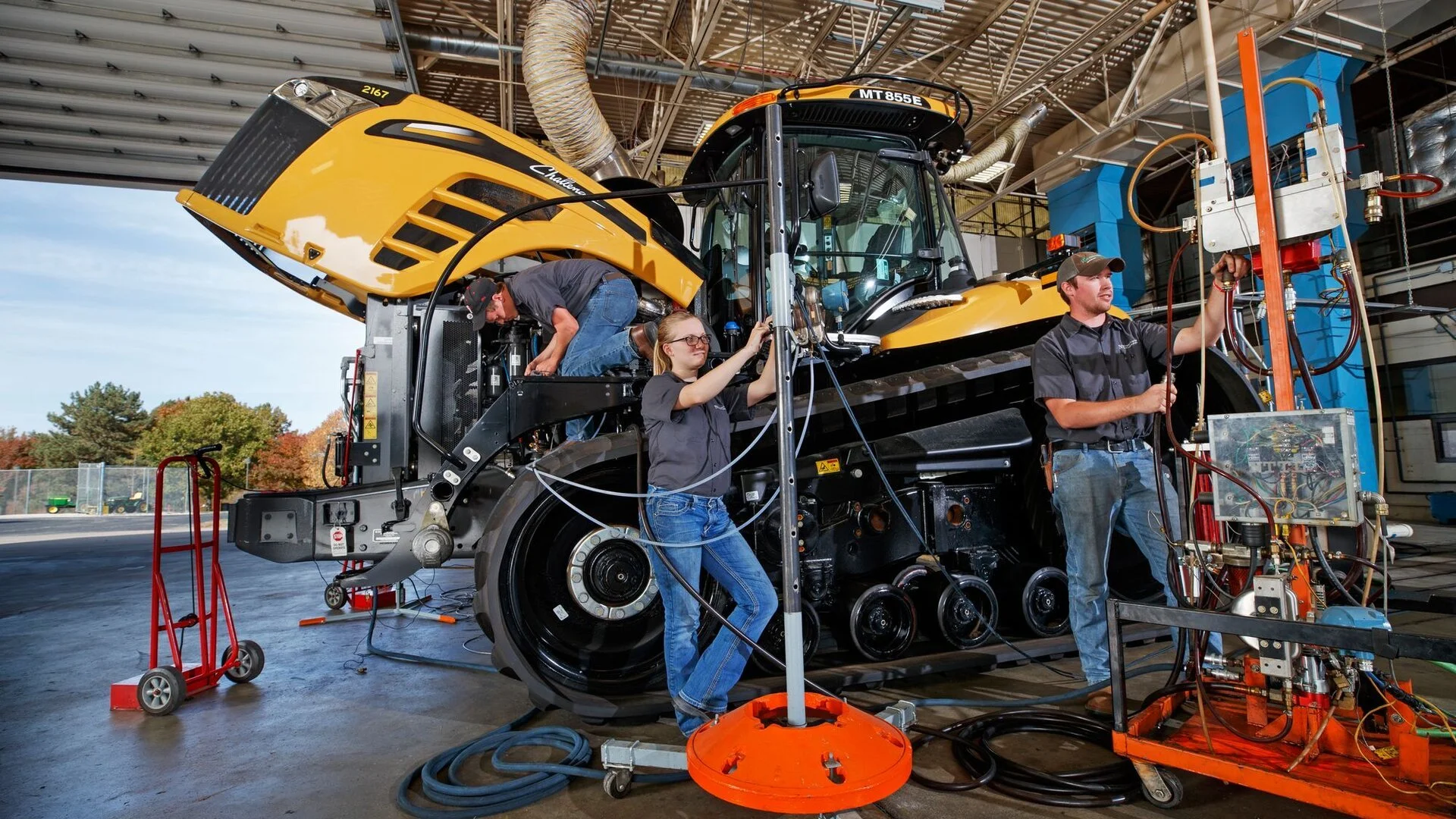 Three workers repair a large yellow construction vehicle inside a factory or workshop. The vehicle is elevated, and the workers are handling various tools and equipment on and around it.