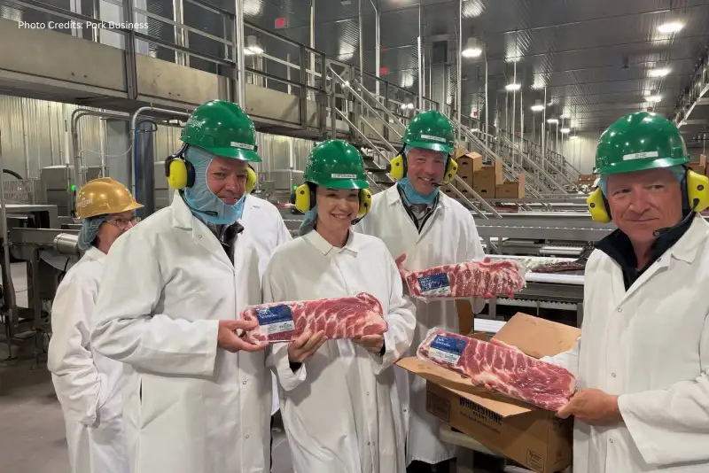 Group of five people in protective clothing and green hard hats holding packages of raw meat inside a meat processing facility.