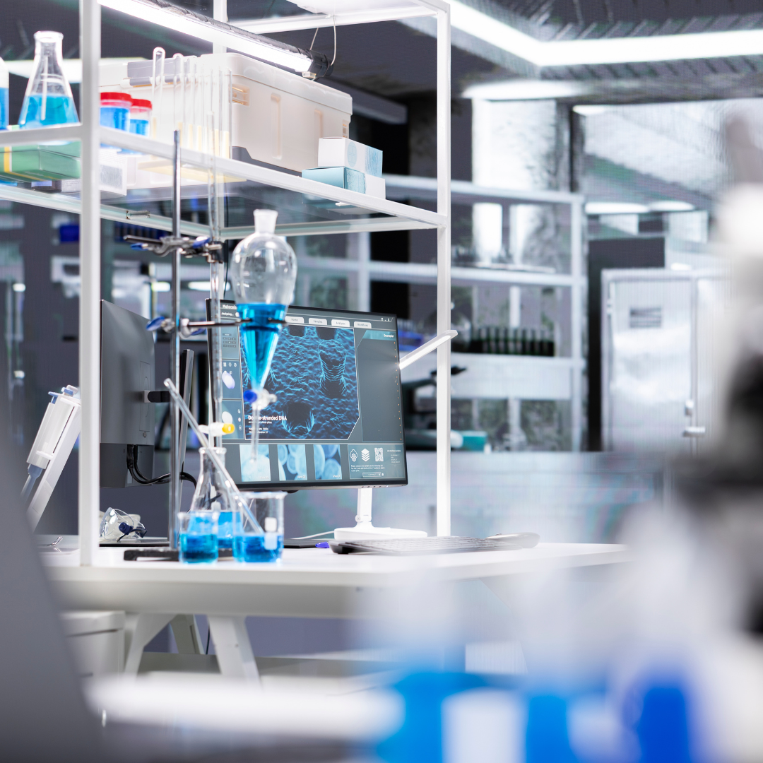 Modern scientific laboratory workspace with glassware, test tubes, a computer monitor displaying DNA or cell structures, and various scientific equipment on a white desk.