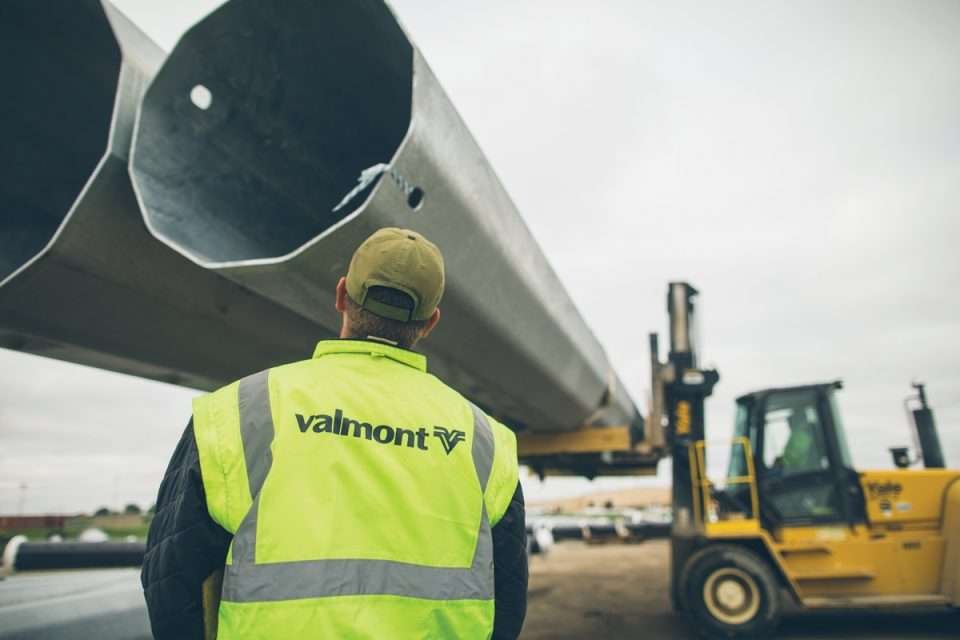 Construction worker wearing a yellow safety vest with 'valmont' on the back, standing in front of large metal utility poles, with a forklift lifting one pole into place at an outdoor construction site under cloudy skies.
